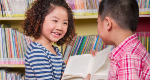 a photo of two Asian children smiling and looking at a book together at a library