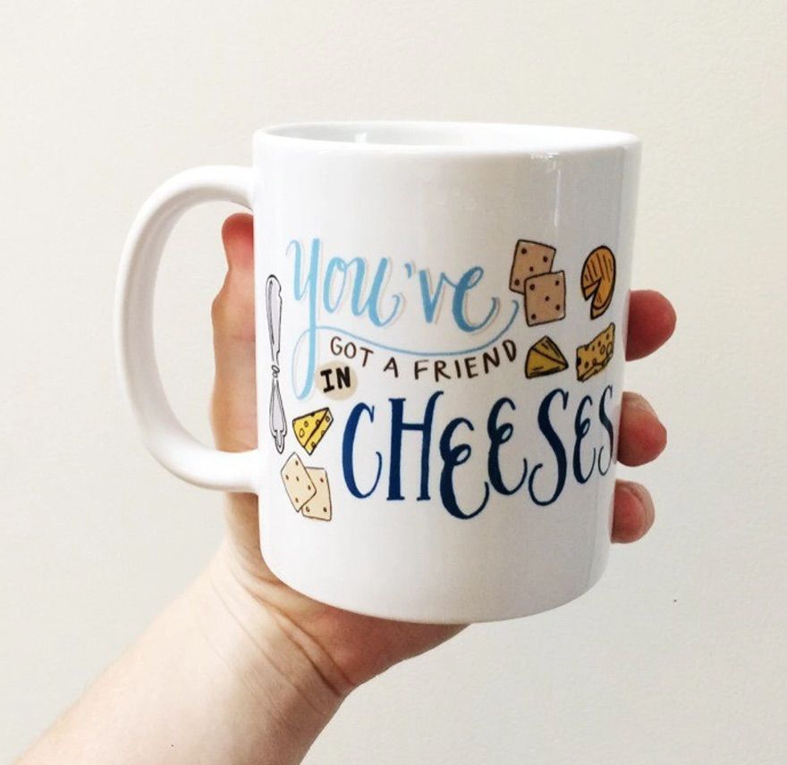 Image of a white mug held by a white hand in front of a cream colored background. On the mug are images of many types of cheese. It reads "you've got a friend in cheeses."
