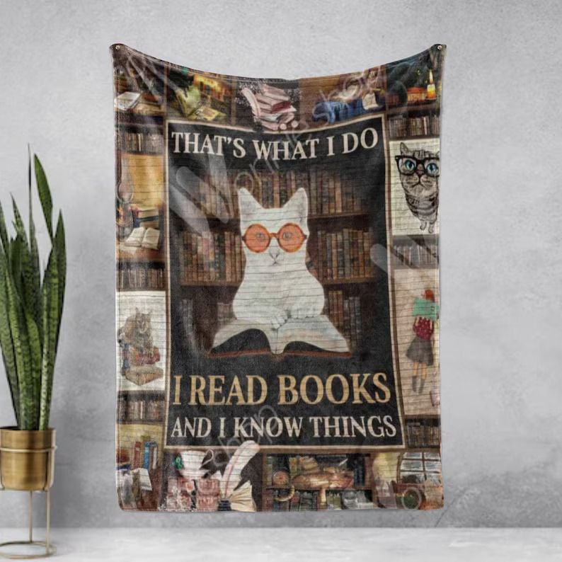 Photo of an open blanket with a myriad of bookish images like cats and books and a girl reading and in the middle a white cat wearing orange classes and holding a book open with a bookshelf filled with books behind it. Above the cat a text reads That's what I do, and below it reads, I read books and I know things
