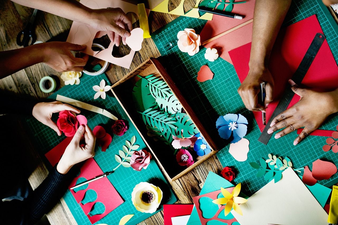 Three people's hands are cutting and folding paper flowers on a table. On the table, there are several other paper flowers in blue and red, as well as some green paper leaves. There are some colourful sheets of paper, and two green cutting boards.