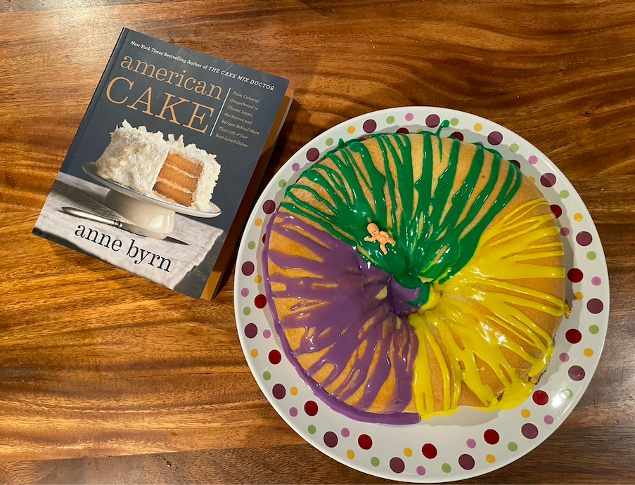 photo of a round king cake with bright green, yellow, and purple drizzle on a dotted serving plate next to the cookbook American Cake