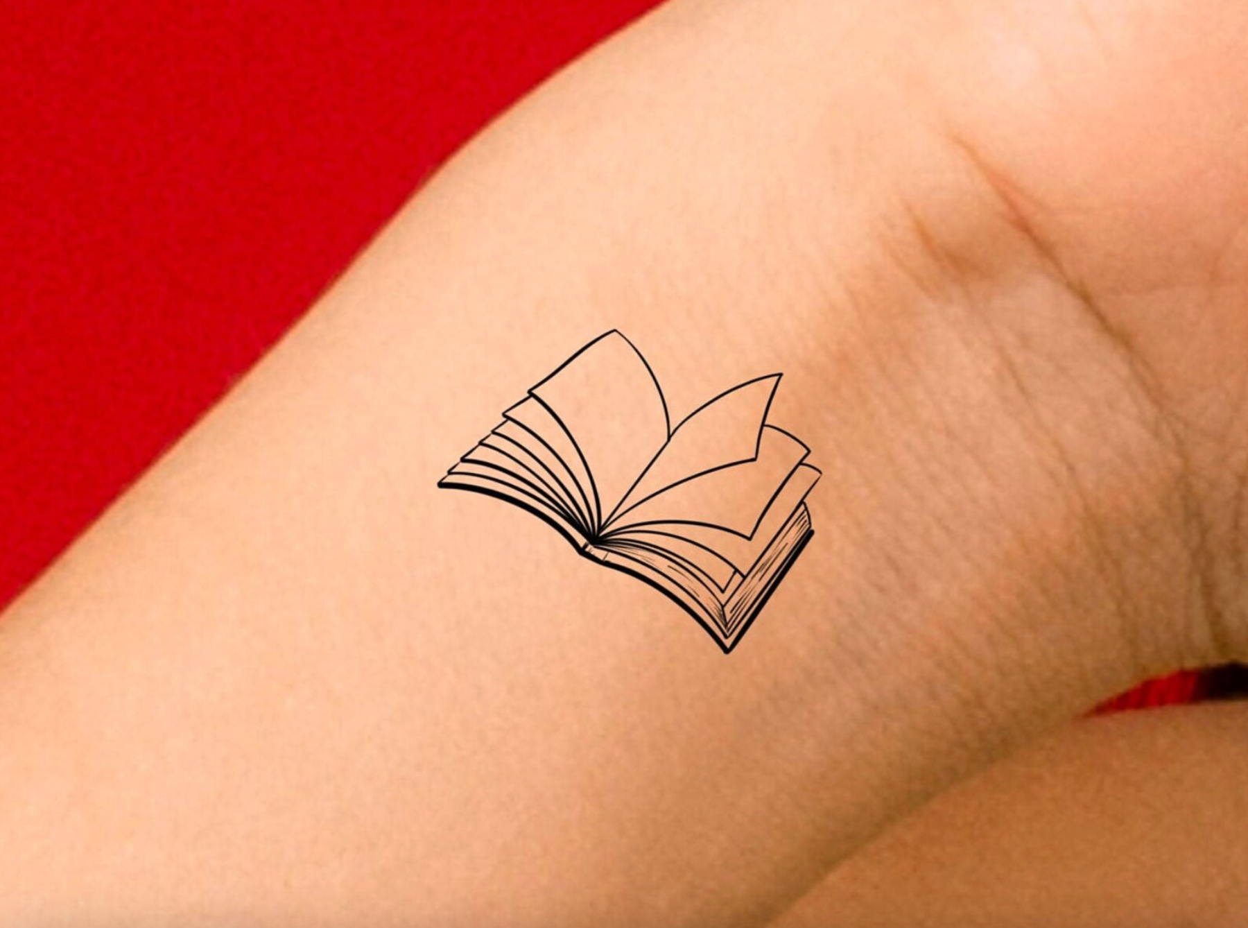 A simple black lined tattoo of an open book on the inside of a person's wrist