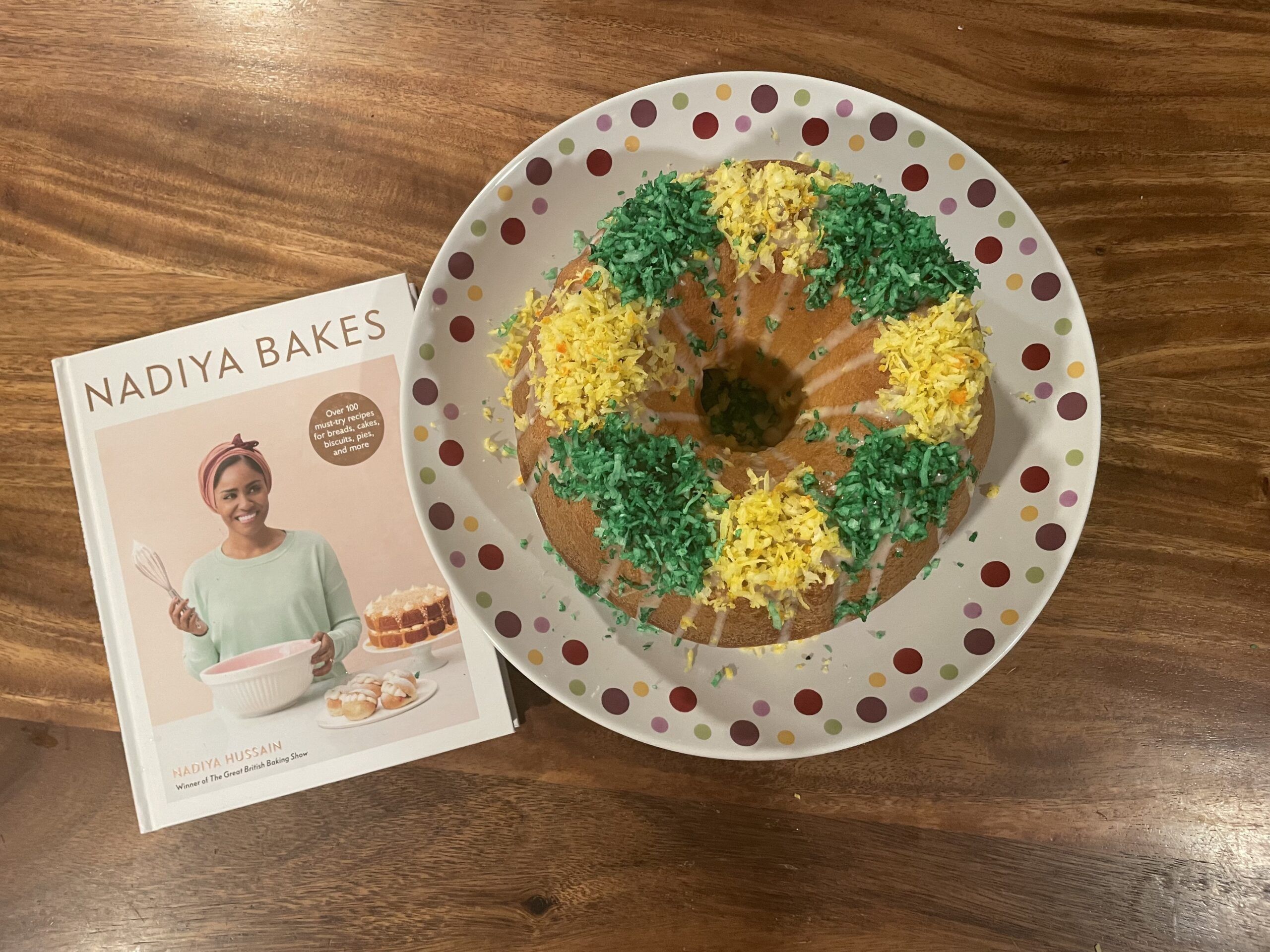 Image of a round king cake topped with green and yellow shredded coconut on a dotted serving plate next to the cookbook Nadiya Bakes