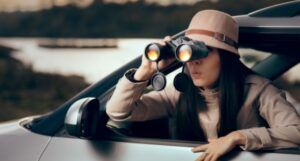 woman with fair skin leaning outside of a car looking curiously through binoculars