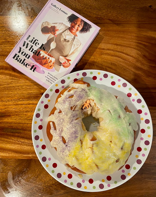 Image of a round king cake topped with thick glaze and purple, green, and yellow sugar on a dotted serving plate next to the cookbook Life is What You Bake It