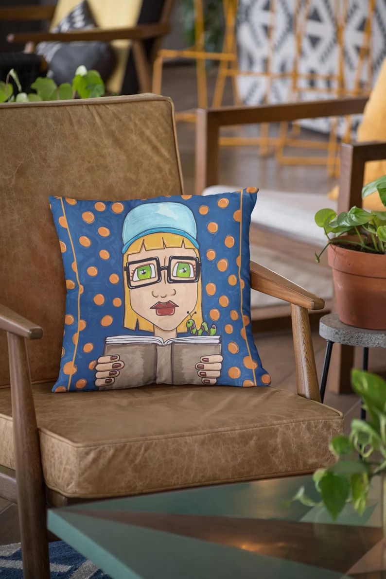 Photo of a pillow with a blue background and yellow polka dots, the drawing of a blond person with green eyes and glasses holding a book with a worm on it.