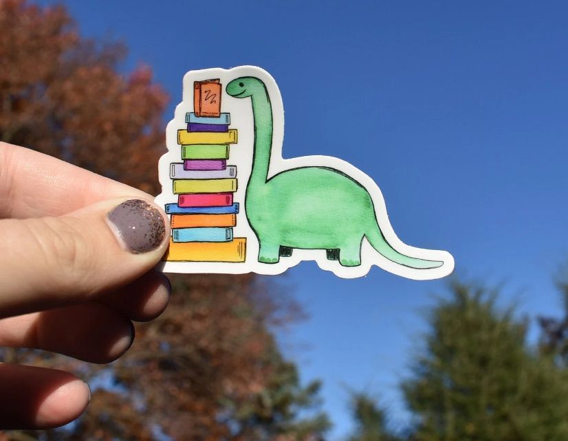 Image of a white hand holding a sticker. The sticker has a green dinosaur with a stack of books. 