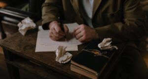 a person in a green blazer sitting at a desk seen from the chest down writing on white paper. On the desk are crumped pieces of paper and a stack of leather-bound notebooks