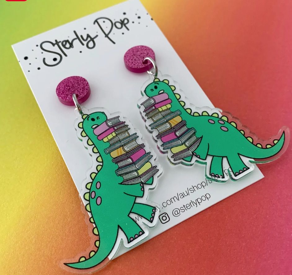 Image of a set of earrings. They are green dinosaurs holding a stack of books. 