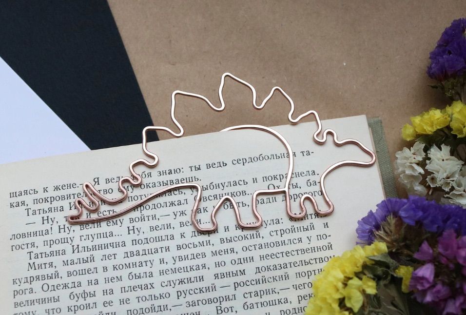 Image of a metal bookmark in the shape of a stegosaurus. It is holding open a book page. 