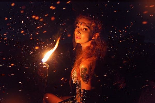 a photo of a person with pale skin and long hair in fantasy dress, holding a torch, looking over their shoulder at the camera