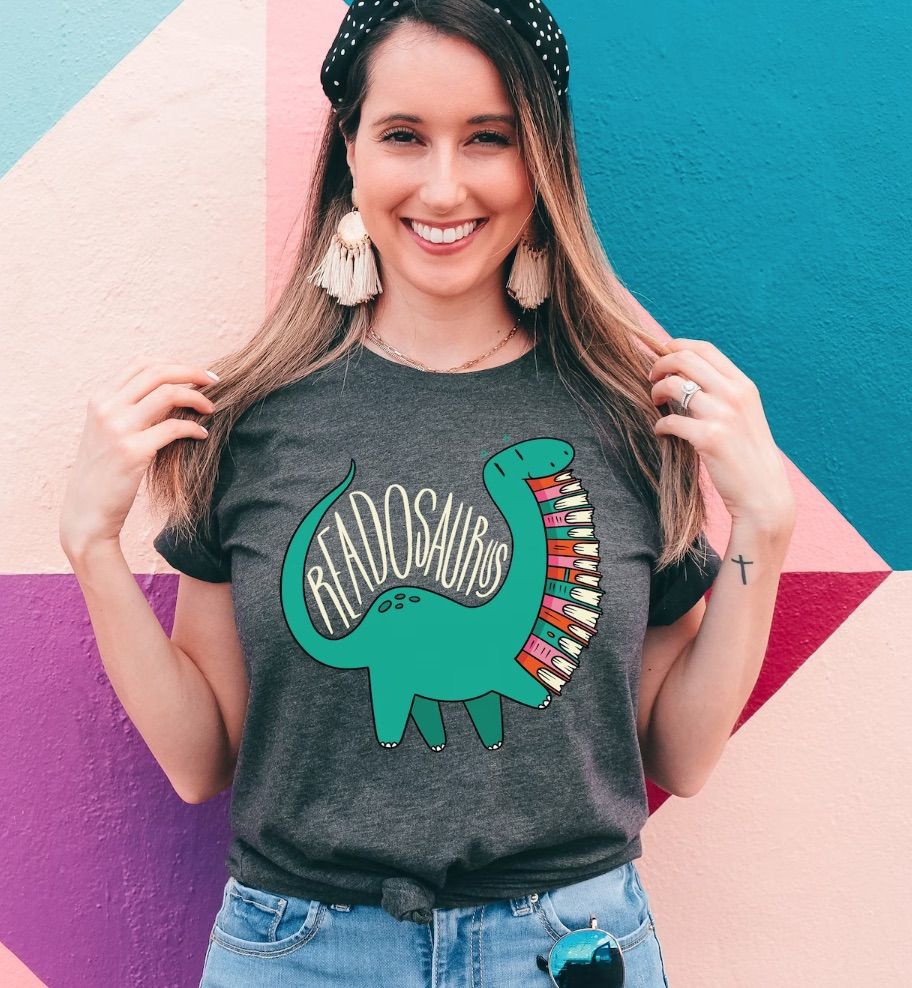 Image of a white person with long blonde hair wearing a grey shirt in front of a colorful wall. The shirt says "readosaurus," and it has a green dinosaur carrying a book stack. 