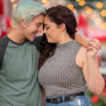 a photo of a sapphic couple holding hands, leaning their foreheads together, and smiling, with lights in the background