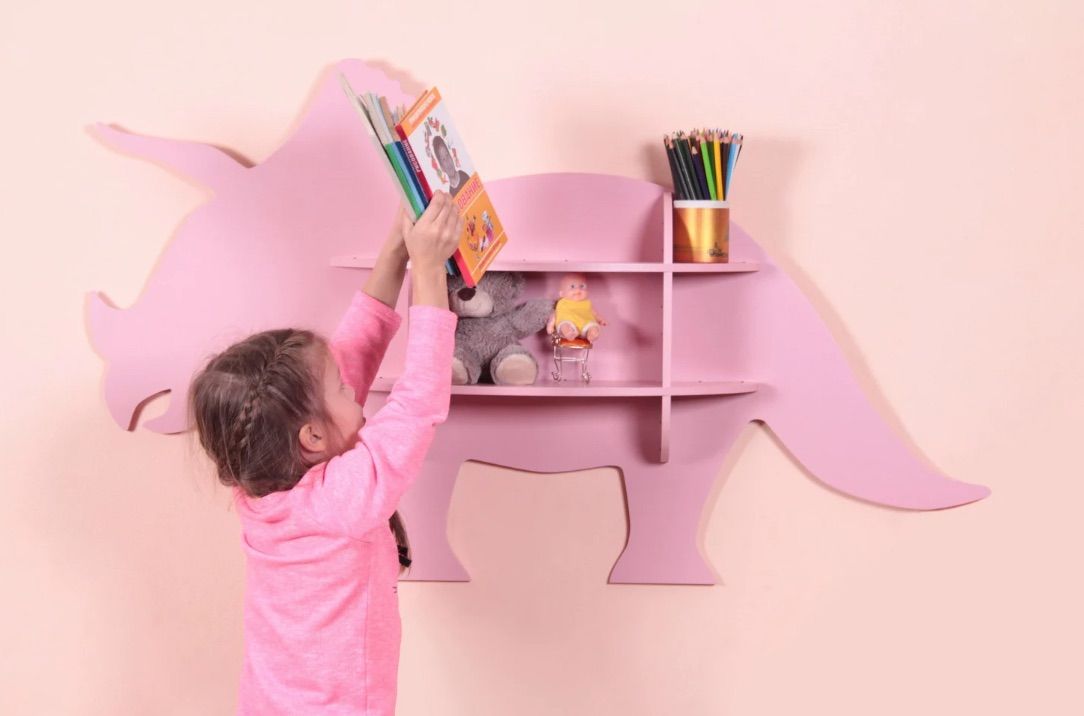Image of a white child shelving books on a shelf in the shape of a triceratops. 