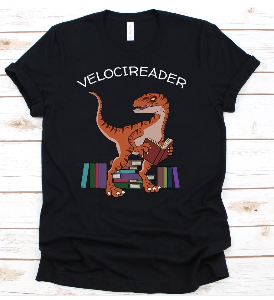 Image of a black shirt with a dinosaur reading a book. The text says "velocireader." 