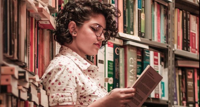 medium-skinned woman leaning on a bookshelf and reading a book