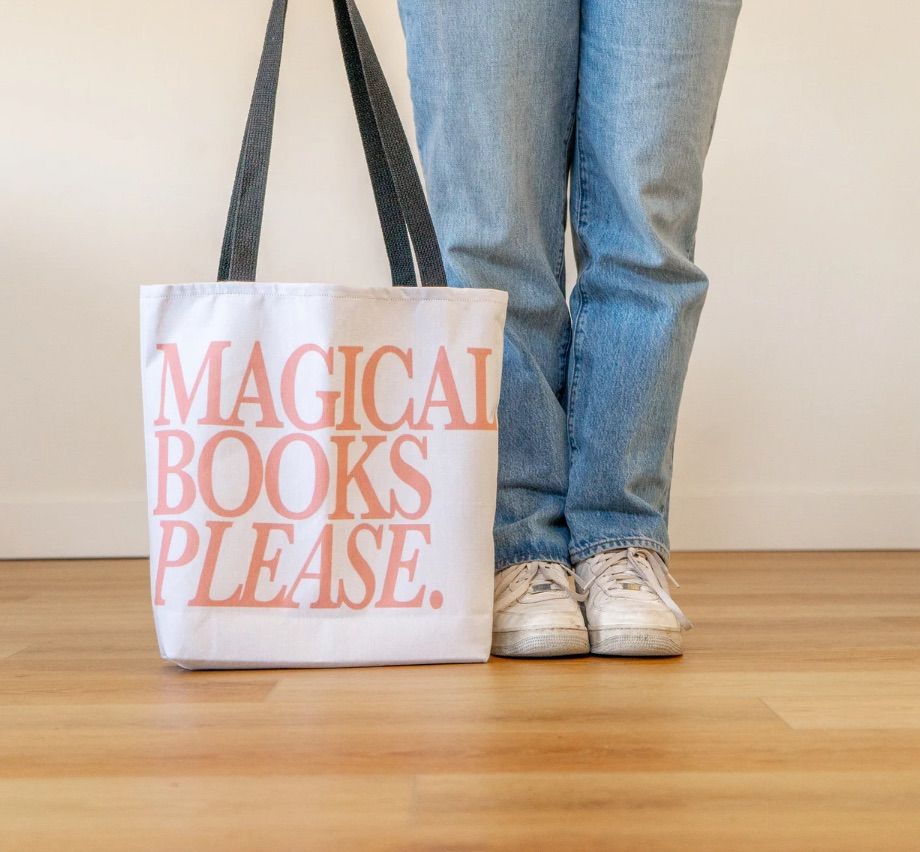 Image of a person holding a white tote bag. The tote says "magical books please" in pink.  