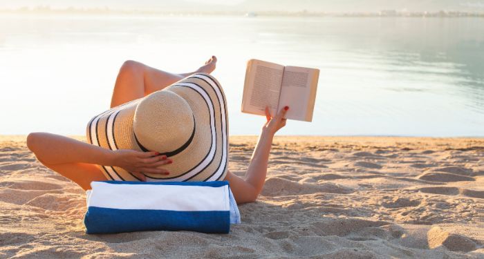 a person in a large straw hat reading a book on the beach as seen from behind