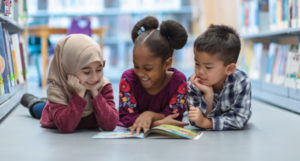 a photo of three kids on the floor reading a book together and smiling