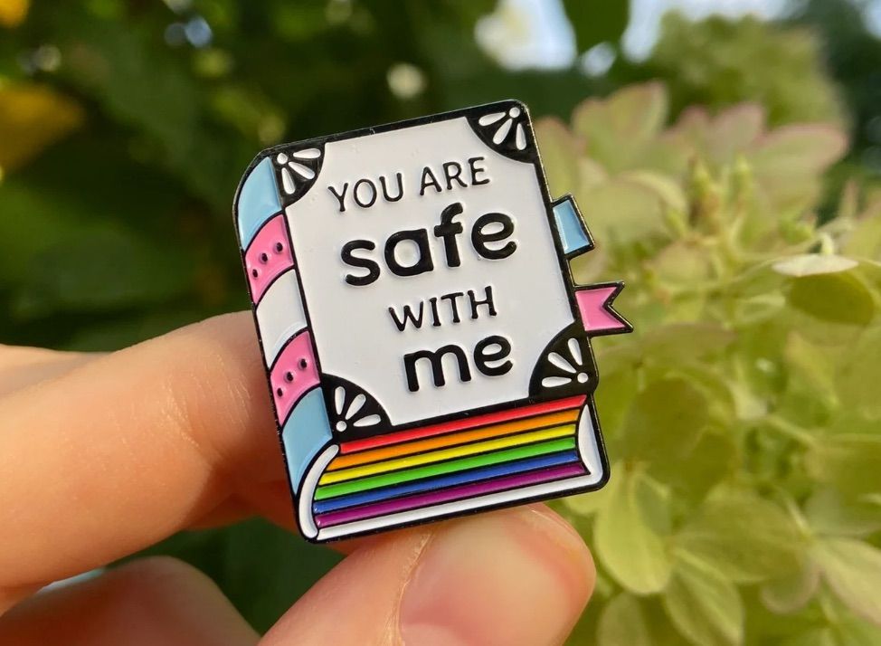 enamel pin of a book with rainbow colored pages that says "you are safe with me"