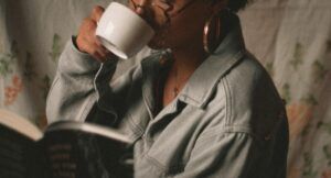 brown-skinned woman with curly hair drinkig out of a white cup while reading a book; she's wearing glasses and a denim jacket