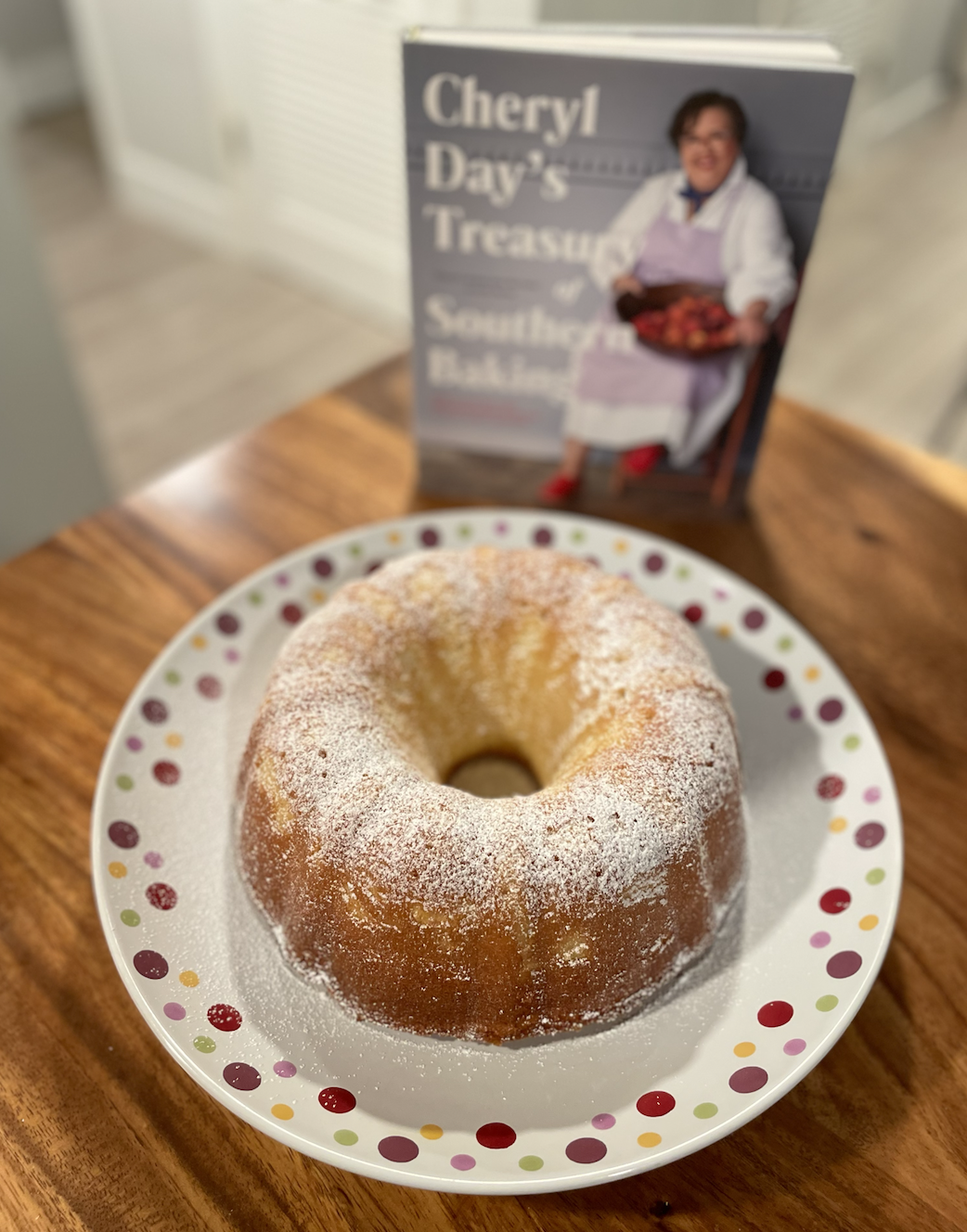 Photo of a light golden Bundt pound cake dusted with powdered sugar on a dotted serving plate on a wooden table next to the cookbook Cheryl Day's Treasury of Southern Baking