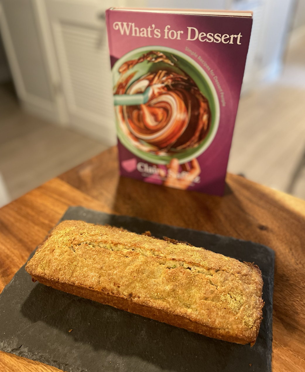 photo of a golden loaf cake on a slate serving board on a wooden table next to the cookbook What's for Dessert