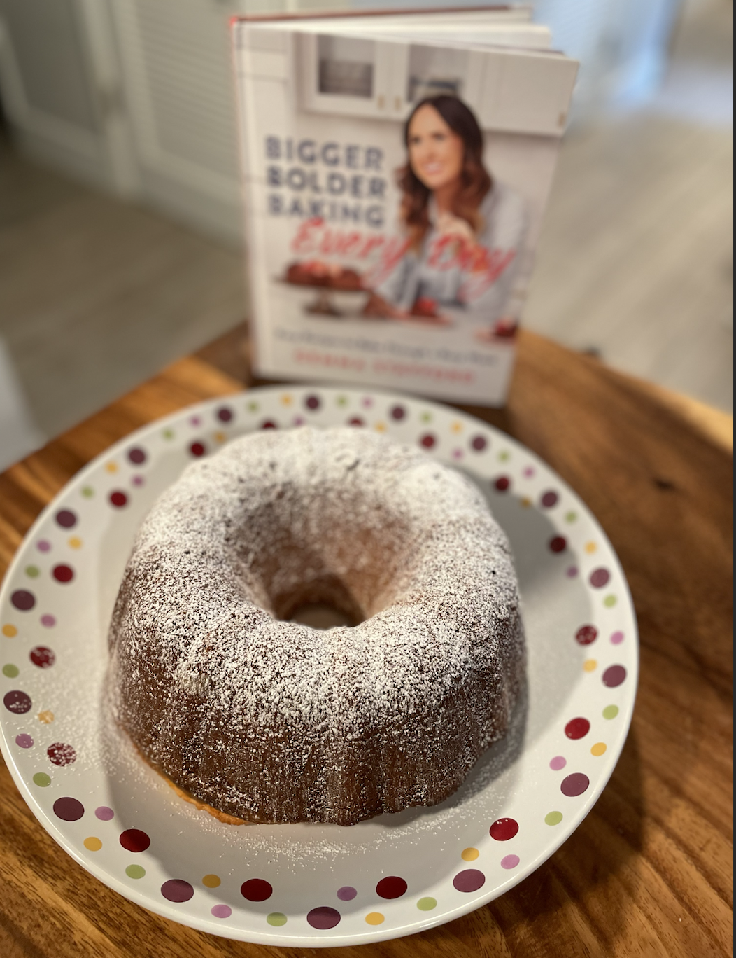 photo of a bundt cake dusted with powdered sugar on a dotted serving platter on a wooden table next to the cookbook Bigger Bolder Baking Every Day