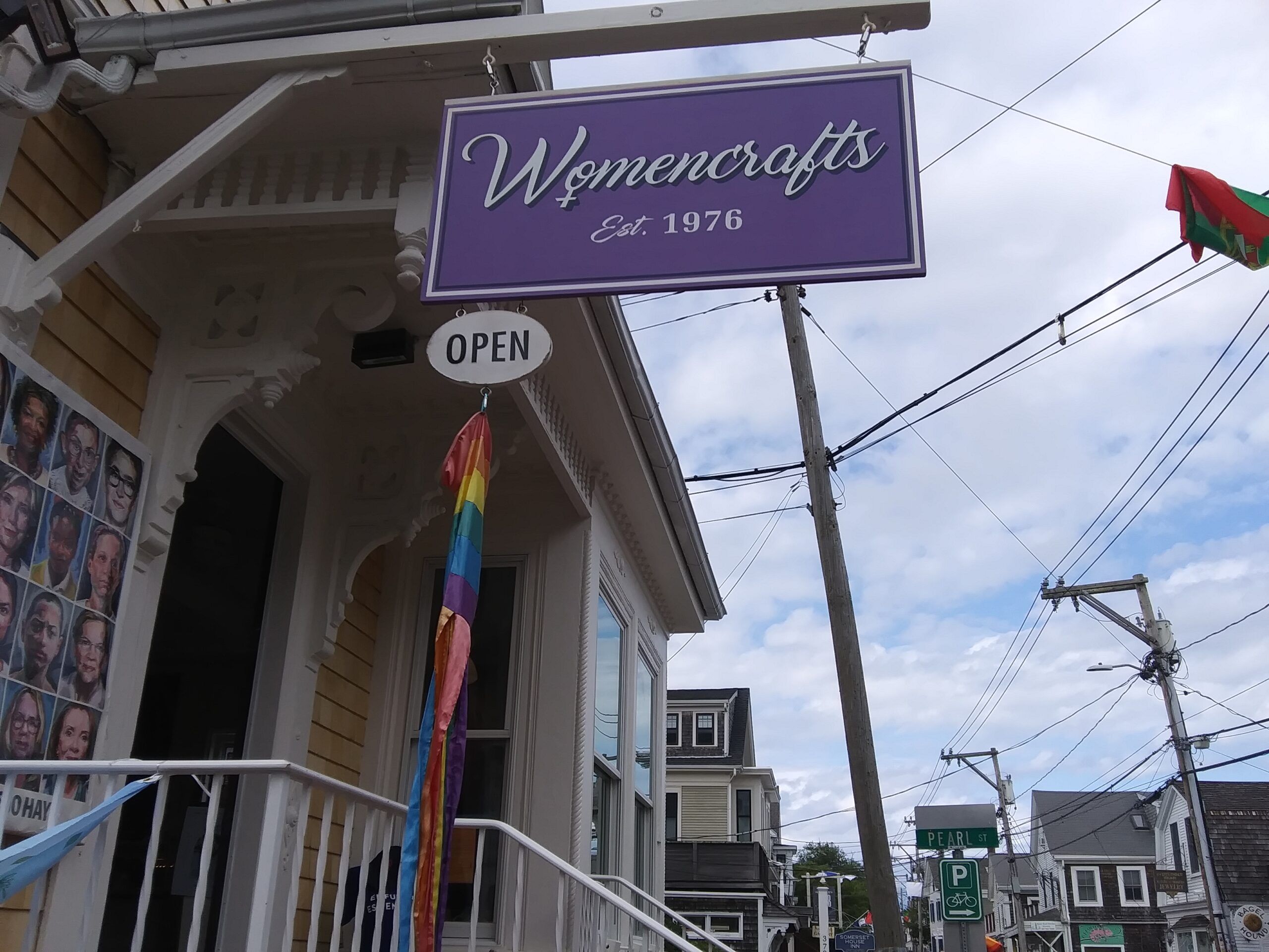 A purple sign hanging over a small yellow-brown building. The sign says Womencrafts in white cursive script