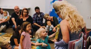 a photo of a Drag Queen Story hour, showing kids looking at a book with a drag queen
