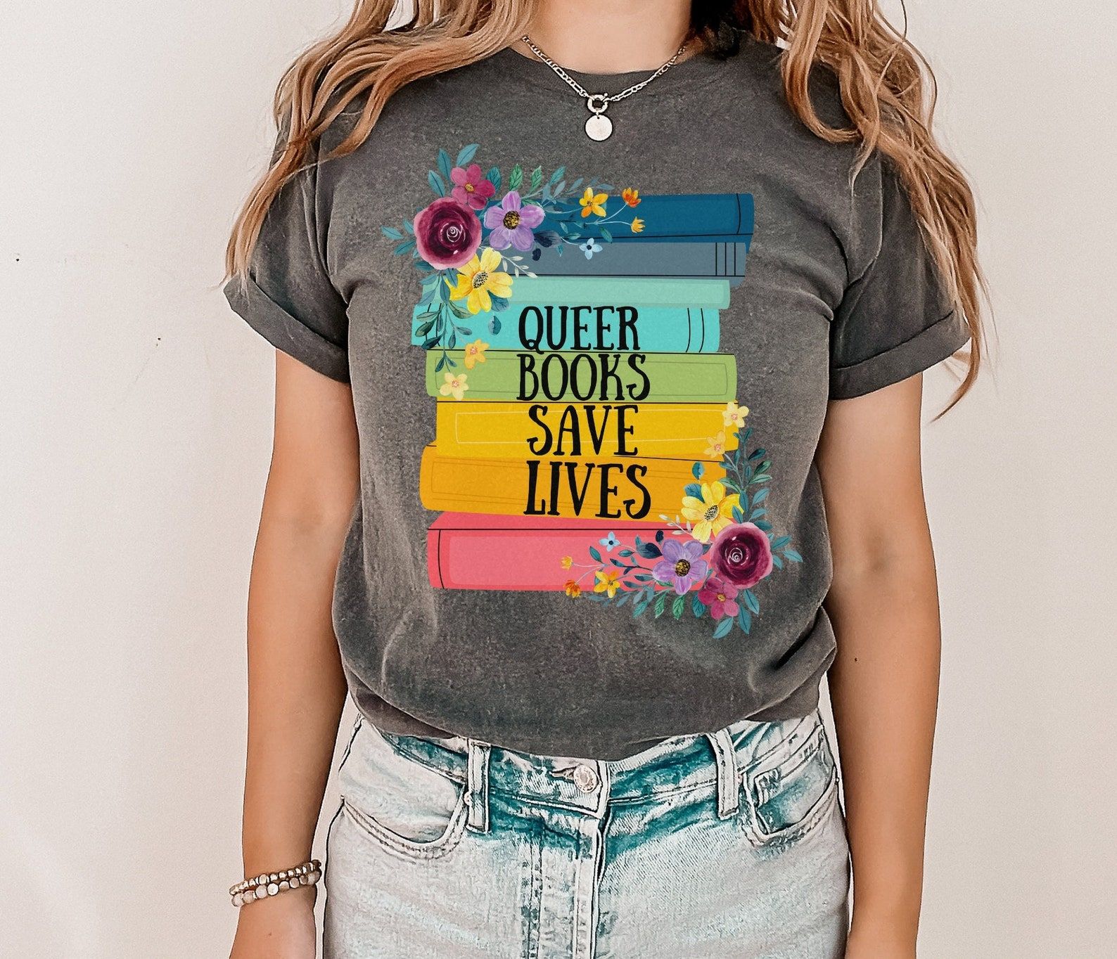 A photo of someone wearing a tee shirt with an illustration of a stack of rainbow colored book spines, florals, and the words "Queer books save lives."