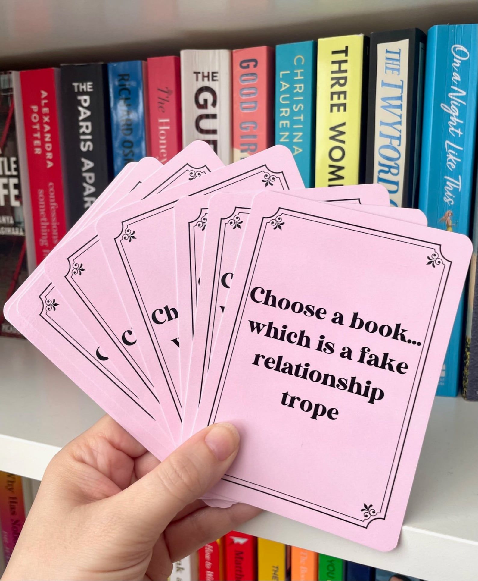 set of pink cards with black writing that prompts various books, such as "choose a book with a fake relationship"