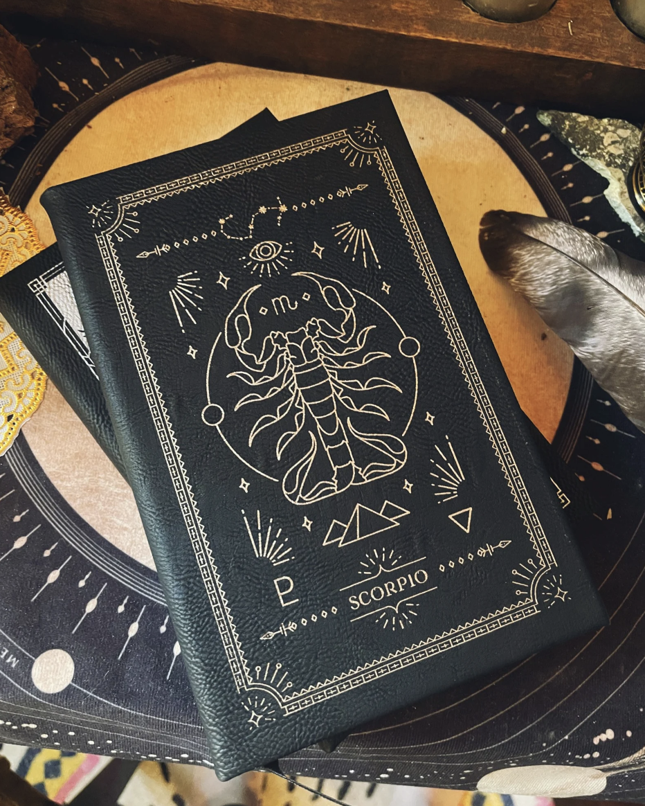 A black faux leather journal with an intricate gold etched illustration inspired by scorpios on the front, resting on a moon backdrop