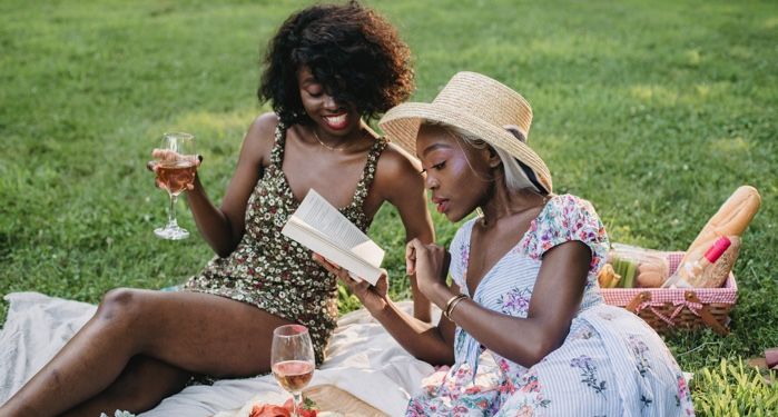 Two dark brown-skinned Black women on a blanket on the grass with books and glasses of wine