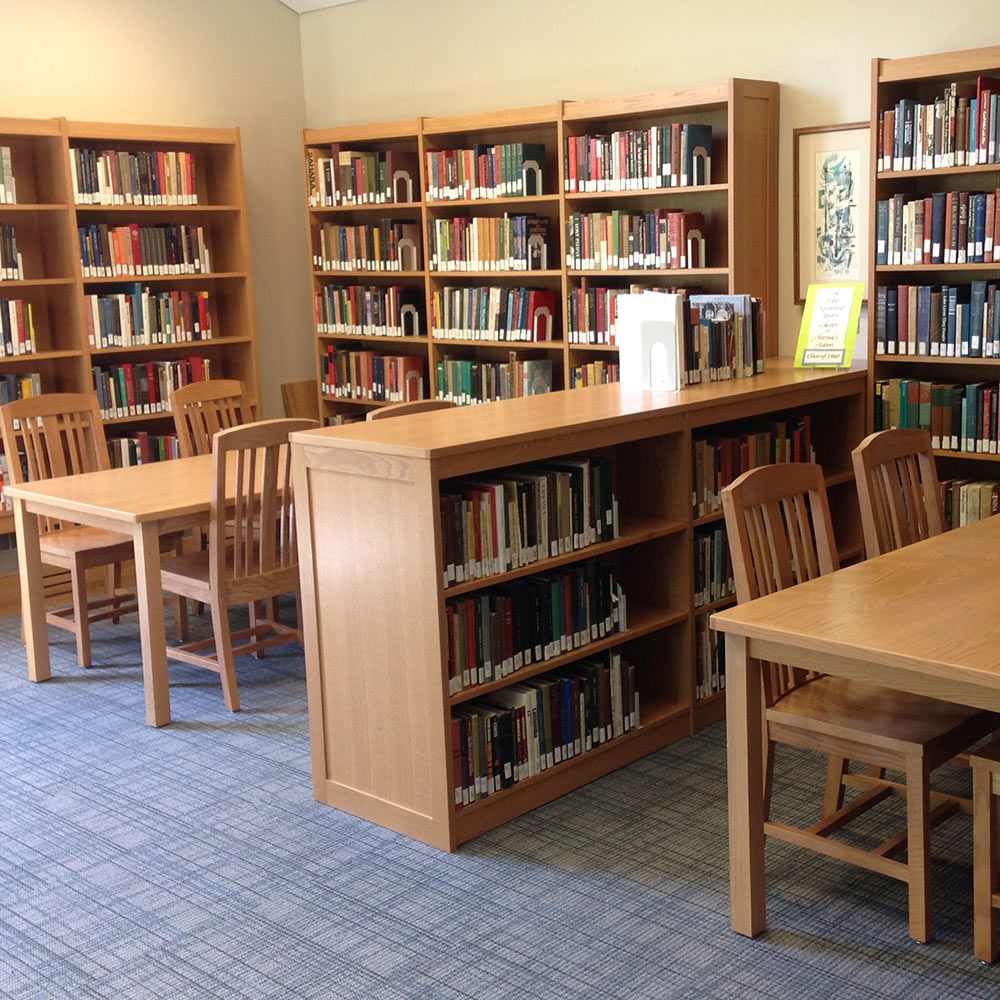 Light wooden tables and chairs surrounded by similar shelving filled with library books. 