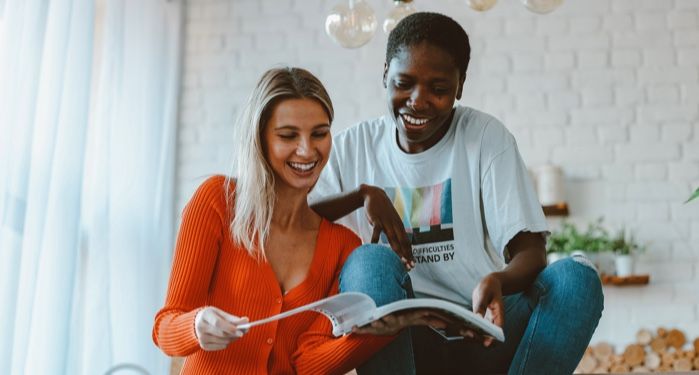 a dark brown-skinned woman with short hair and a fair-skinned white woman laughing at some reading material