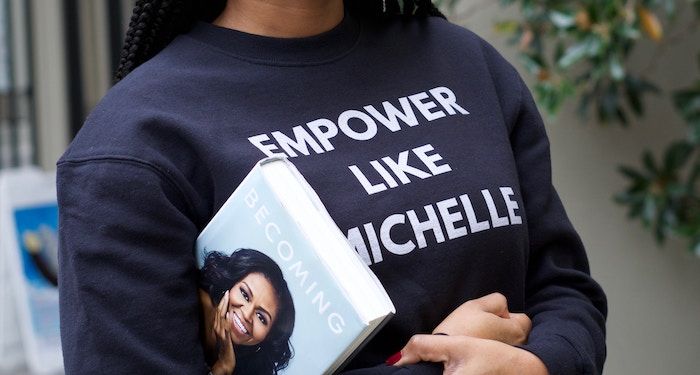 a photo of a Black woman wearing a shirt saying "Empower like Michelle" with Michelle Obama's memoir tucked under one arm