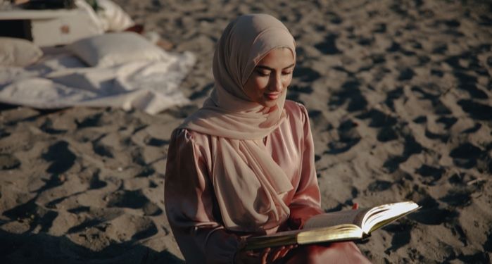 medium light skinned Muslim woman wearing a light pink hijab and reading on the beach