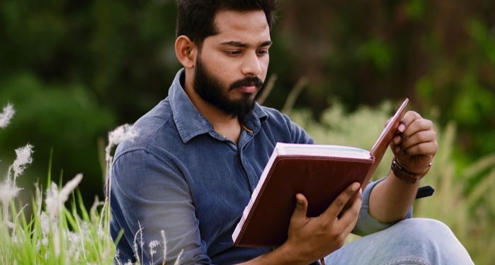 medium skin-toned South Asian man reading a book in the grass near flowers