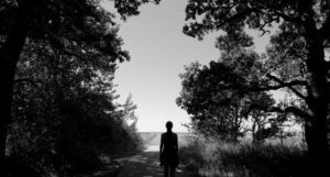black and white image of a person standing in a dirt road with grass and trees on either side