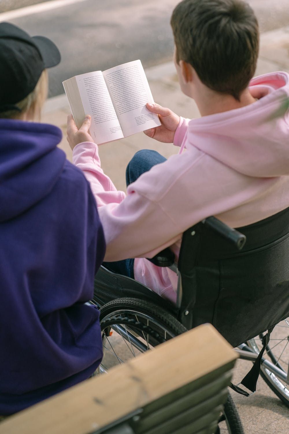 Seen from behind, a white person in a wheelchair, with short brown hair and a pink hoodie, is holding a book and appears to be reading aloud to another white person with blond hair, a black baseball cap and a dark blue hoodie
