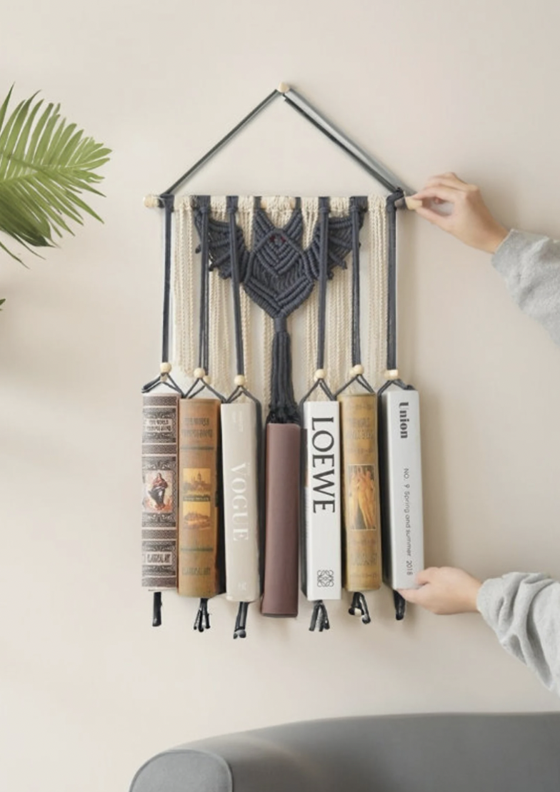A hanging bookshelf made of taupe yarn with a gray crocheted bat at the top hangs on a wall holding up seven books