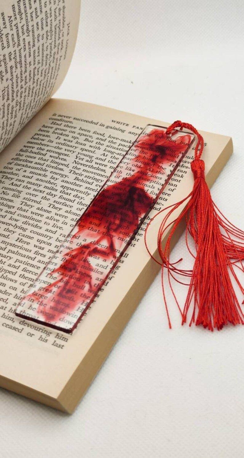 Photo of a slightly open book with a bookmark on top of the pages. The bookmark is made of clear resin and seems to have blood running through it, it also has a string at the top. 