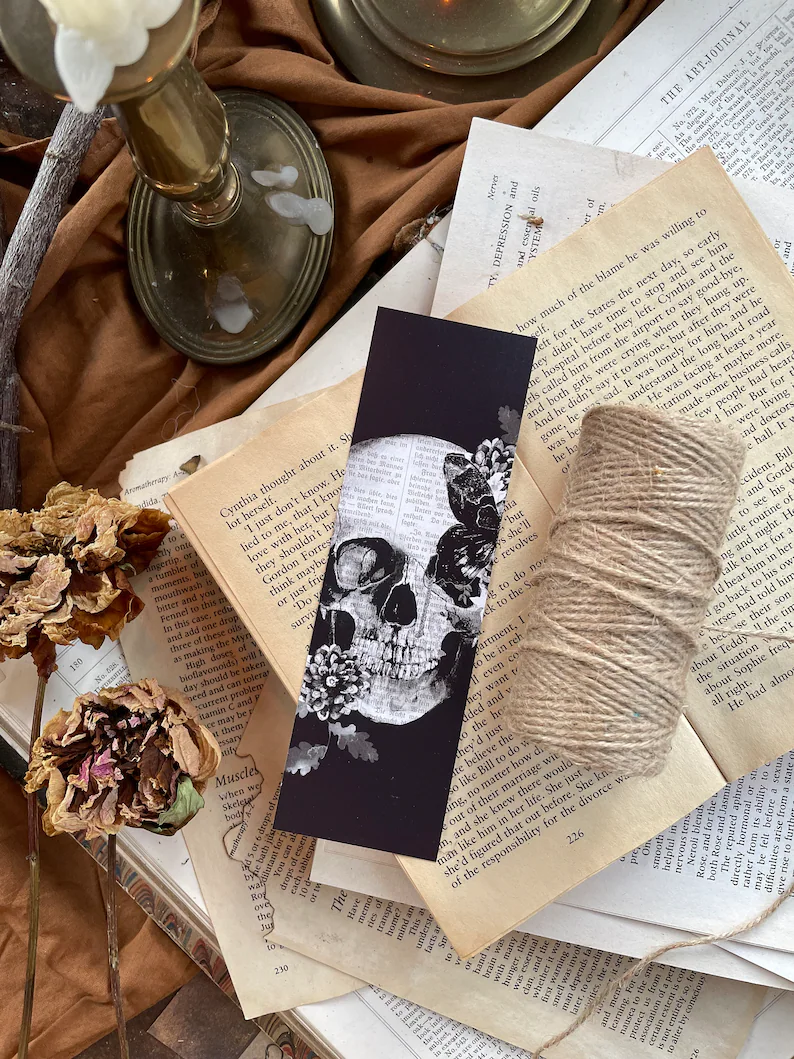 Photo of a bookmark with a skull framed by some flowers placed on an open book on a table filled with string and dried flowers and loose pages. 