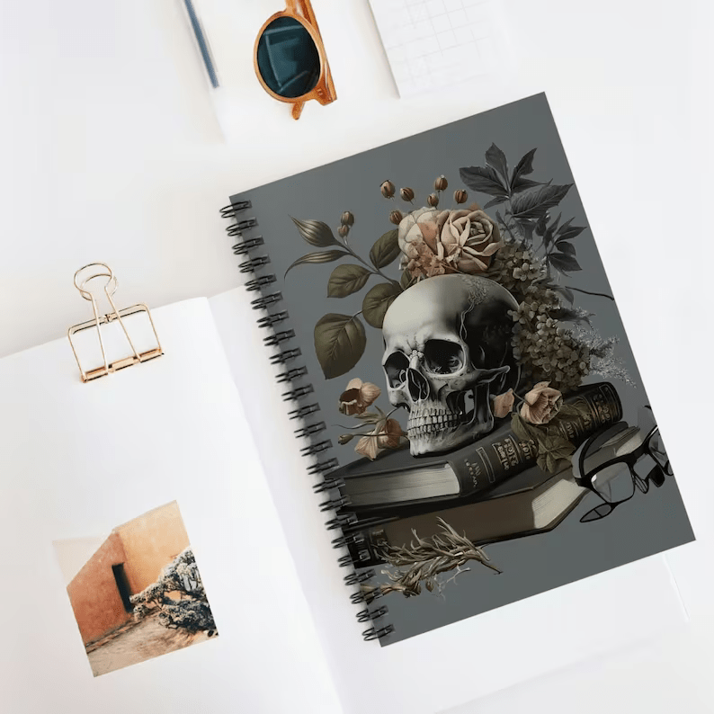 Photo of a notebook with a spiral spine placed on a table with some paper next to it, and some pens and sunglasses above it. The cover shows a skull placed on top of some books with flowers behind it. 