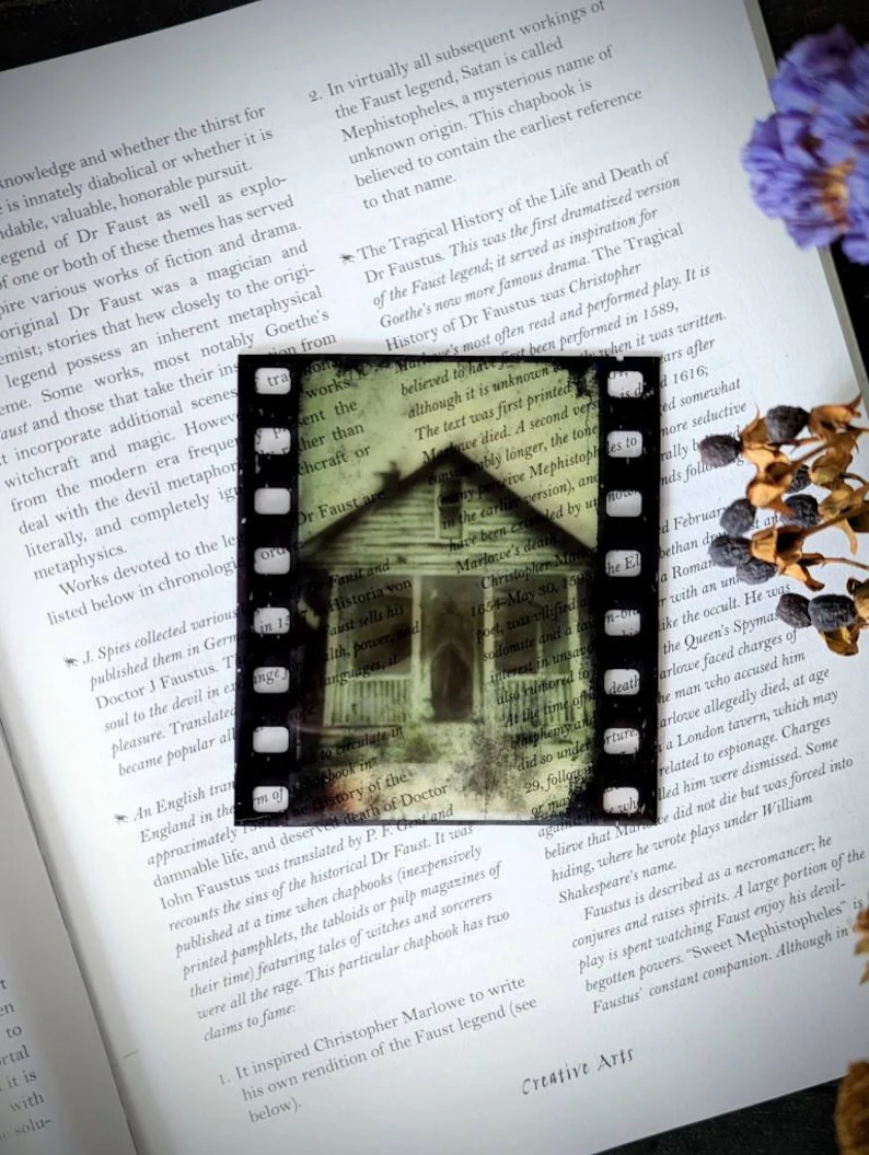 Photo of an open book and a clear bookmark placed on the pages. The bookmark resembles an old film with the image of a haunted house.