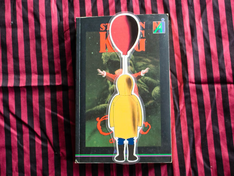 Photo of a bookmark with the shape of a boy wearing a yellow rain jacket, carrying a red balloon placed on top of a copy of IT by Stephen King. 