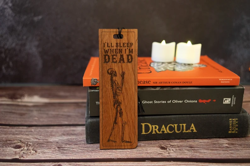 Photo of a wooden bookmark with a skeleton and the text "I'll sleep when I'm dead" engraved in it standing again the spines of three books with two candles on top.