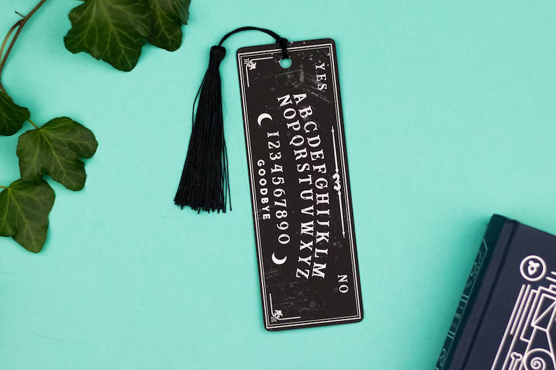 Photo of a bookmark mimicking a Ouija Board placed on a green background with a book on the right and some leaves on the left.
