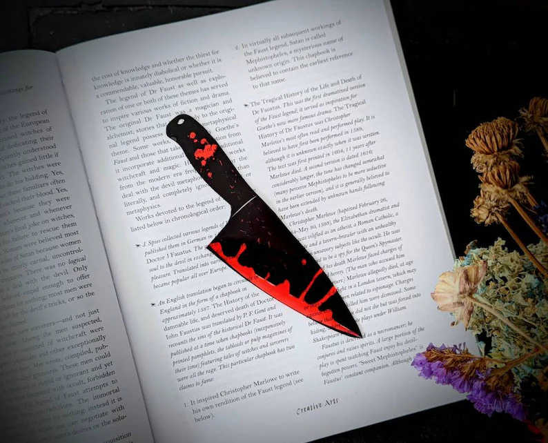 Photo of a bookmark shaped like a bloody knife placed on top of an open book. 